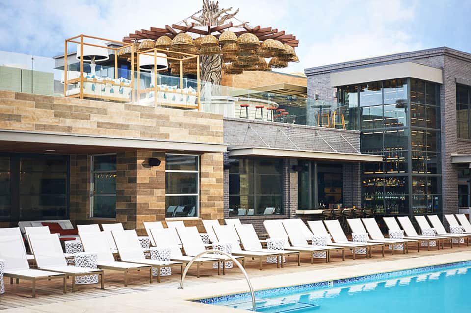 Resort pool deck with lounge chairs, modern building, and decorative overhead structure against cloudy sky