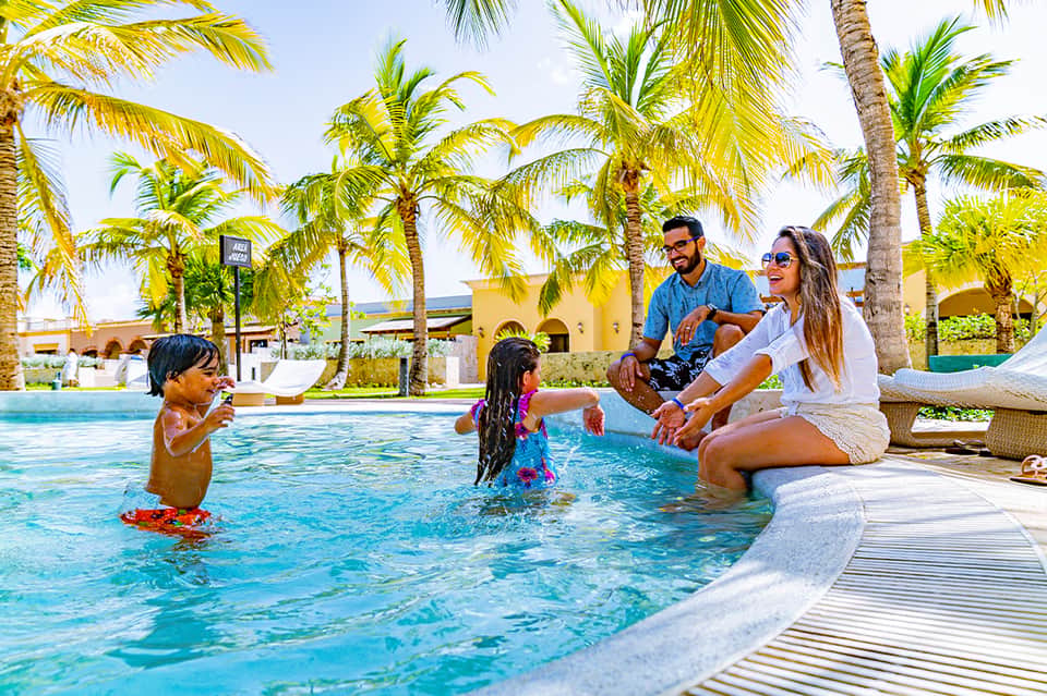 Family enjoying resort pool with children playing in water under palm trees and yellow buildings