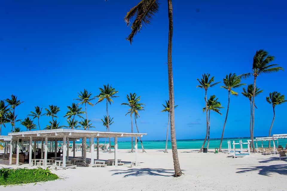 White beach pavilion with turquoise ocean, palm trees, white loungers, and clear blue sky