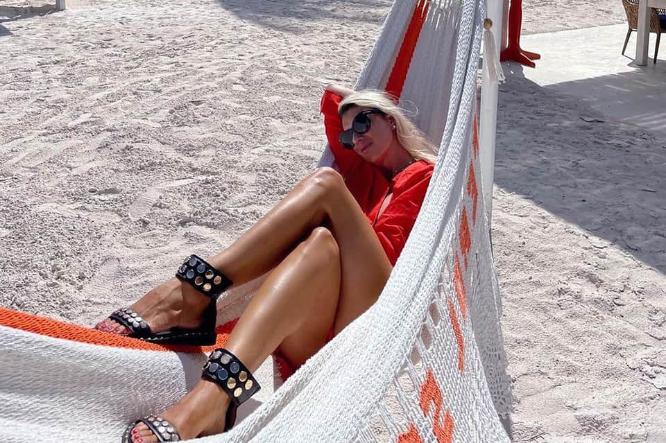 Woman relaxing in red beach lounger on sandy shore wearing sunglasses and studded sandals