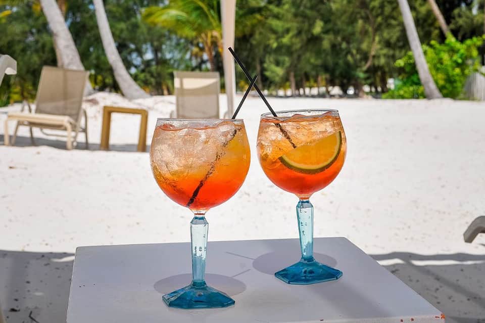 Two tropical cocktails with ice and citrus garnish on white table with beach and palm trees in background