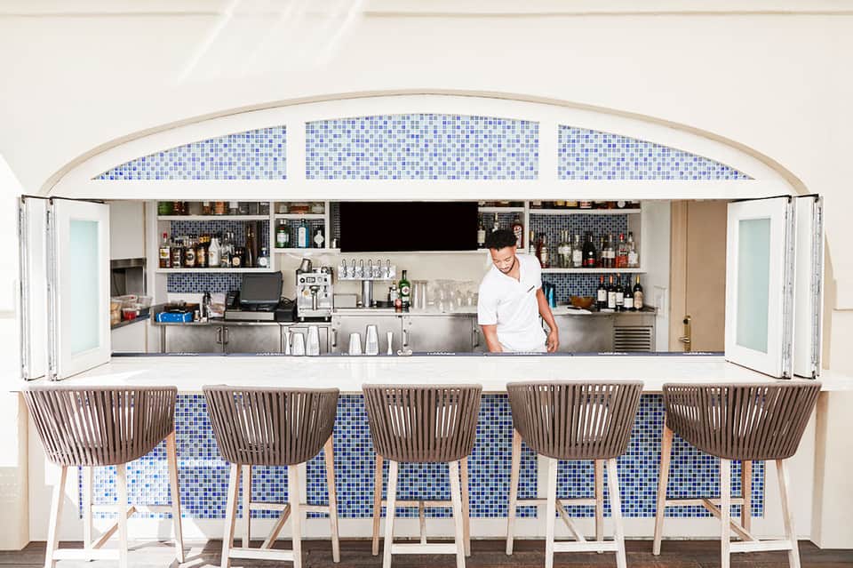 Modern bar with blue tile backsplash, stainless steel counter, and five woven chairs facing bartender in white shirt