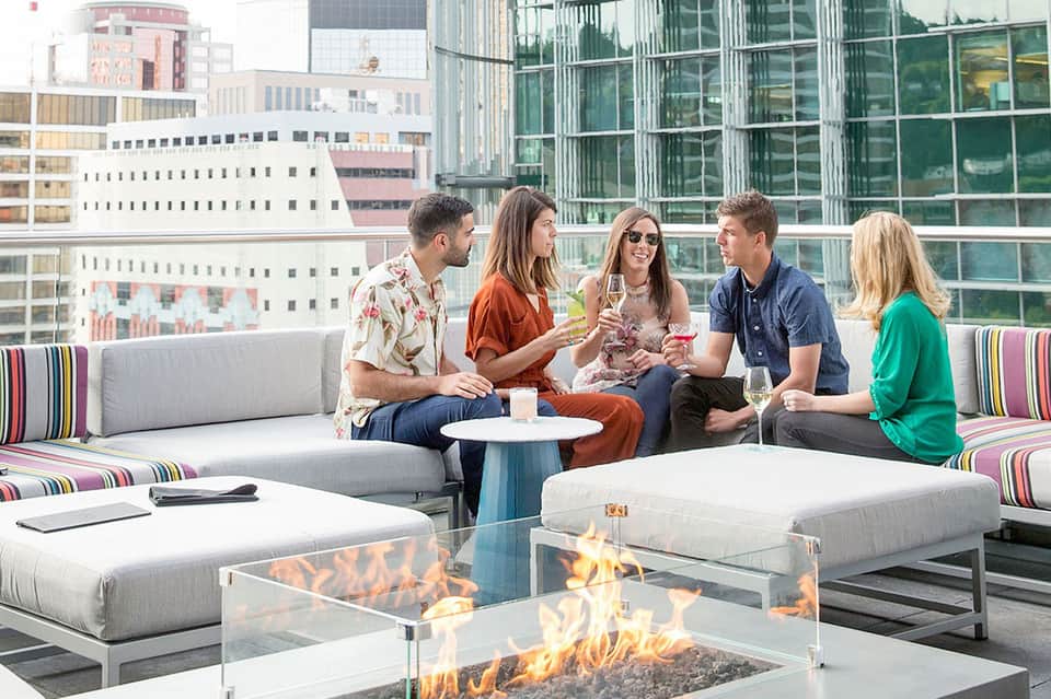 Group of five people socializing on rooftop lounge with firepit, modern seating, and city skyline