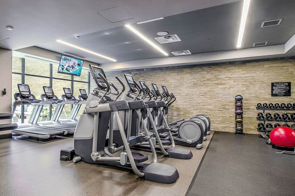 Modern fitness center with rows of elliptical machines, treadmills, free weights, and brick accent wall