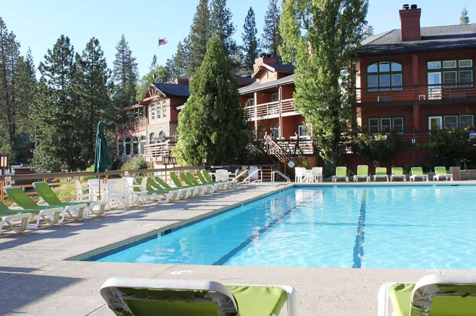 Resort swimming pool with green loungers, alpine lodge buildings, and forest backdrop