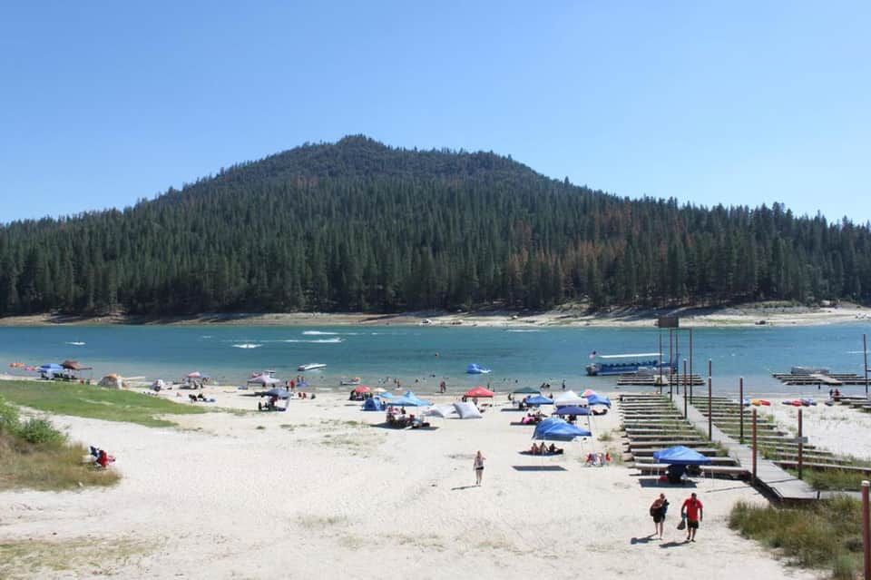 Mountain lake beach with sandy shore, umbrellas, swimmers, docks, and pine forest backdrop