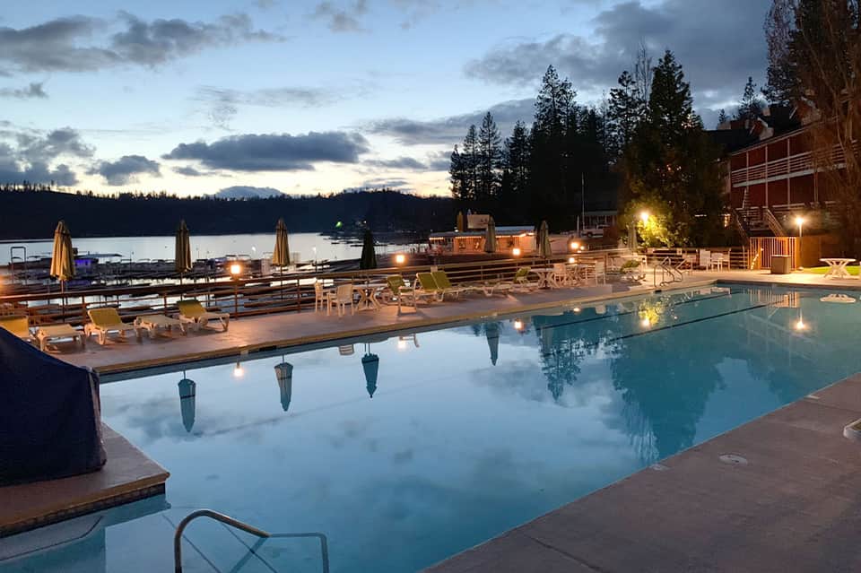 Resort pool overlooking mountain lake at dusk with lit umbrellas and forest backdrop