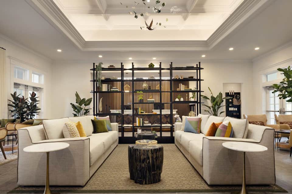 Elegant hotel lounge with cream sofas, black shelving unit, potted plants, and coffered ceiling
