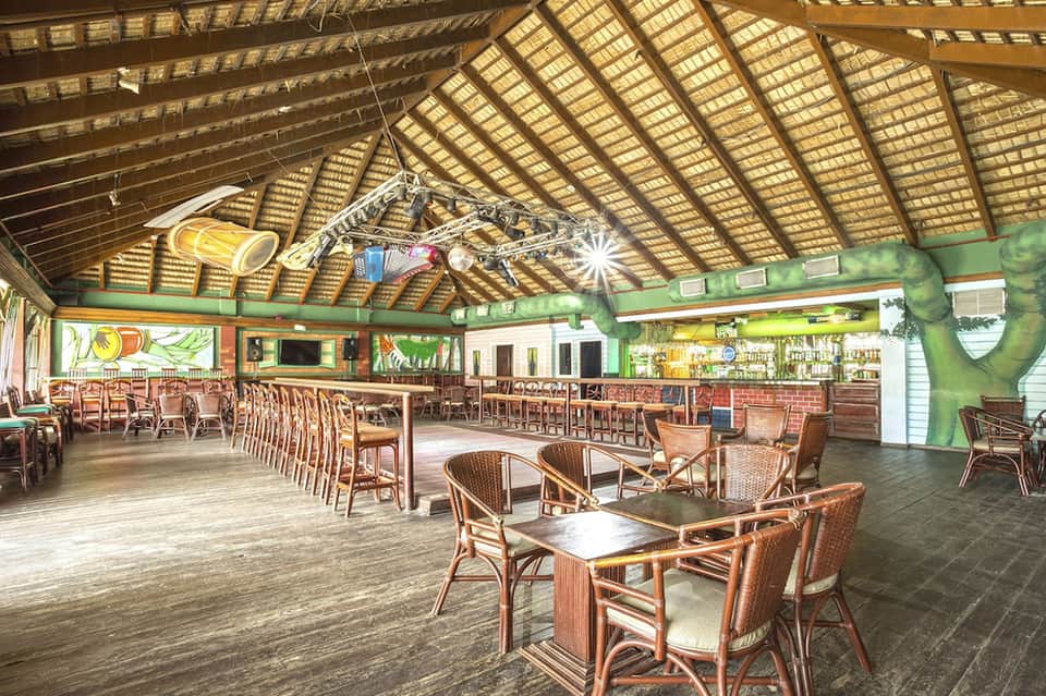 Open-air tiki bar with thatched roof, wooden furniture, bar seating, and tropical décor lighting