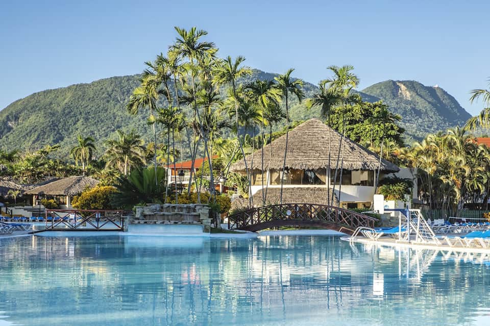 Resort pool with thatched-roof pavilion, mountain backdrop, and tropical landscaping