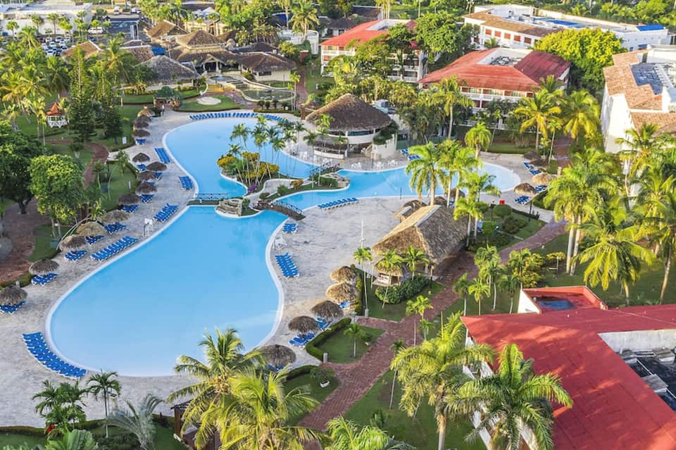 Aerial view of resort's curved blue pool complex surrounded by palm trees and buildings