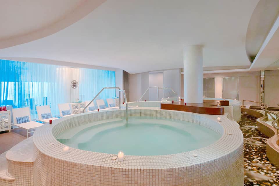 Indoor spa with large circular hot tub, white tile surround, lounge chairs, and floor-to-ceiling windows
