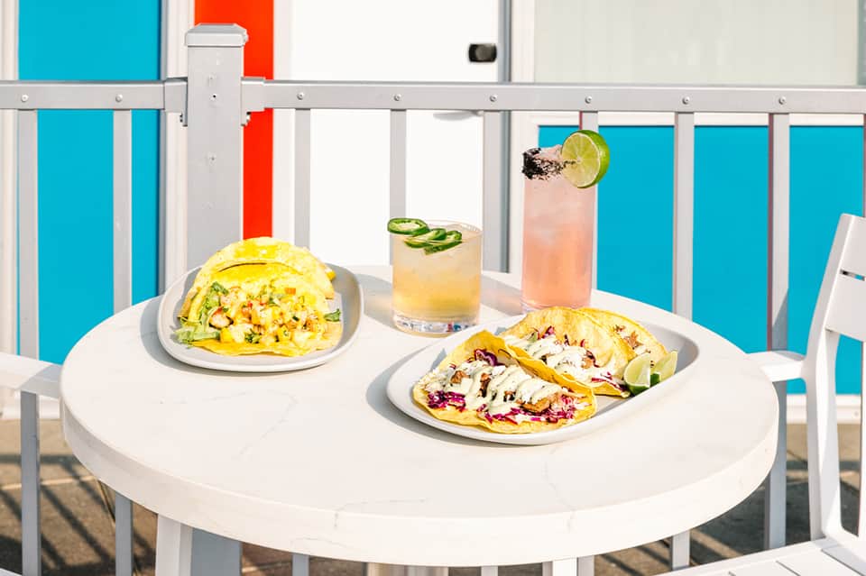 Colorful tacos and cocktails on white table with turquoise pool visible behind railing