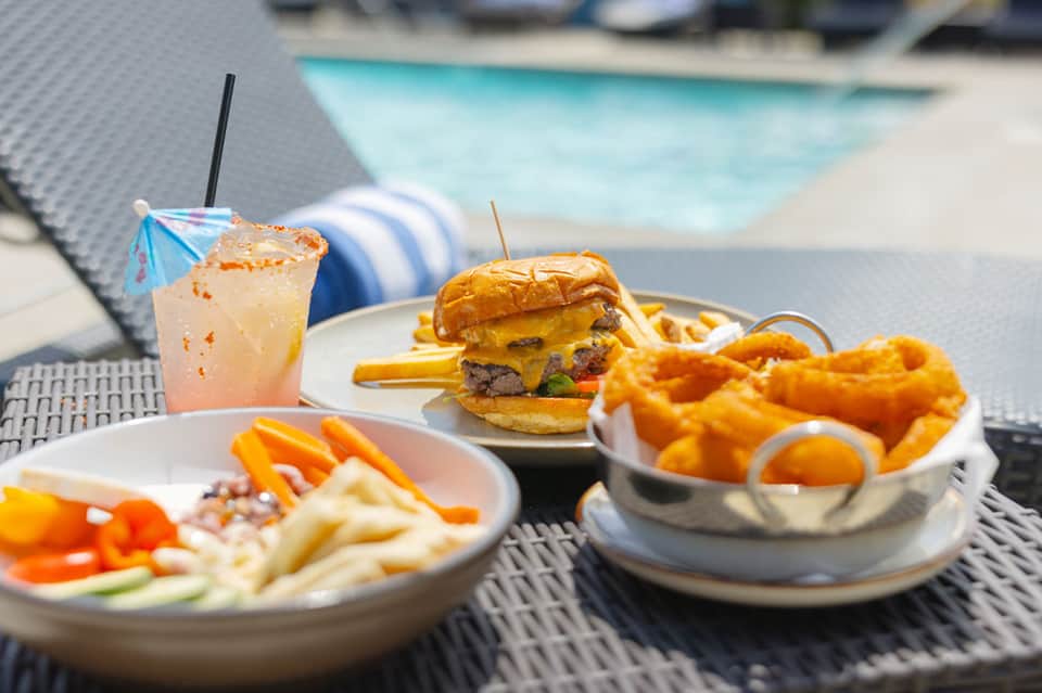 Poolside lunch setting with gourmet burger, onion rings, cocktail, and striped lounge chair