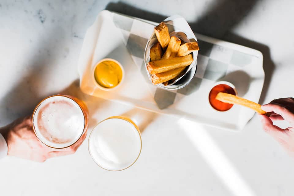 Overhead view of appetizers, fries, and beers on white table in bright natural light