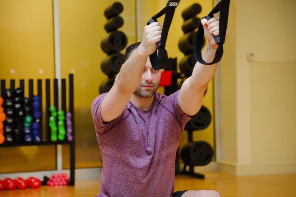 Man in purple shirt performing suspension training exercise with black TRX straps in fitness gym