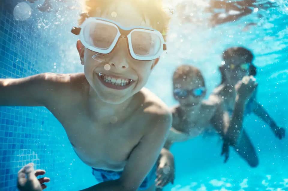 Child wearing goggles swimming underwater in blue pool with other swimmers in background