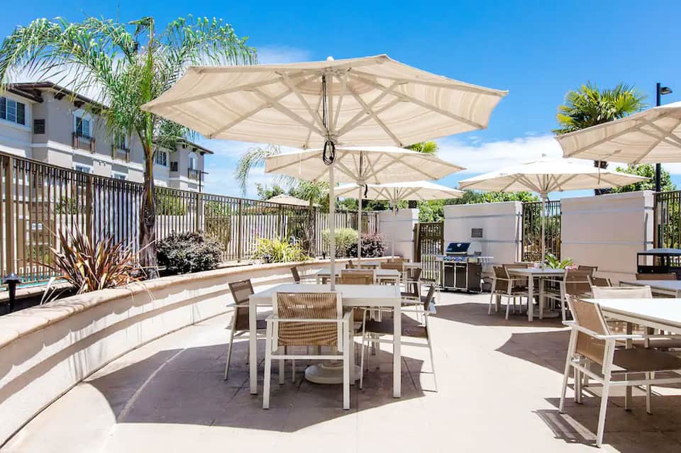 Resort patio with large white umbrellas, dining tables, white fence, and tropical palm trees