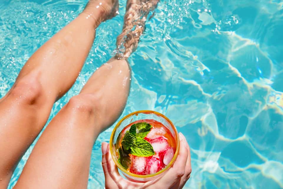 Person holding colorful tropical cocktail by pool with legs in turquoise water