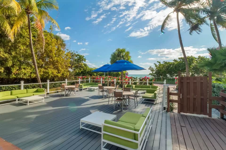 Oceanfront deck with green lounge chairs, blue umbrella, palm trees, and water views