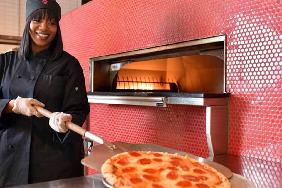 Chef in black uniform presenting fresh pizza from red-wall oven in modern kitchen