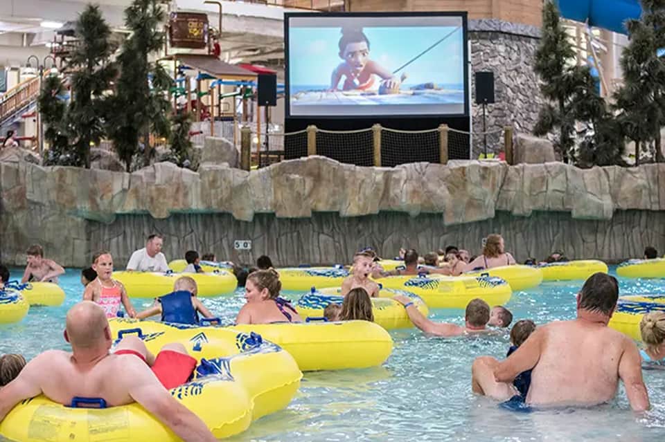 Indoor water park with children on yellow floats, large screen, and stone features