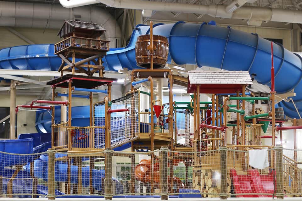Indoor water park with blue tube slides, wooden play structures, and safety netting in warehouse-style facility