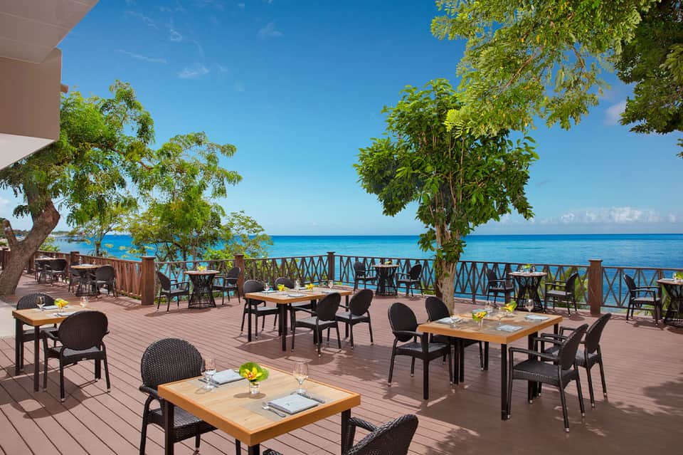 Oceanside restaurant deck with wooden tables, chairs, and tropical tree shade near the water