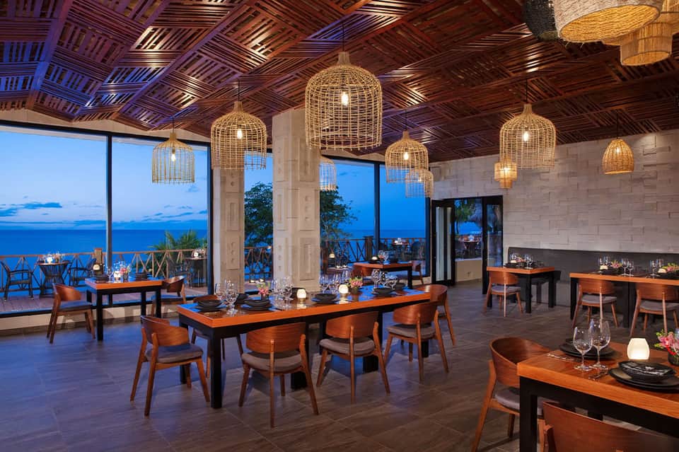 Oceanview fine dining restaurant with wooden tables, woven pendant lights and sunset sea backdrop
