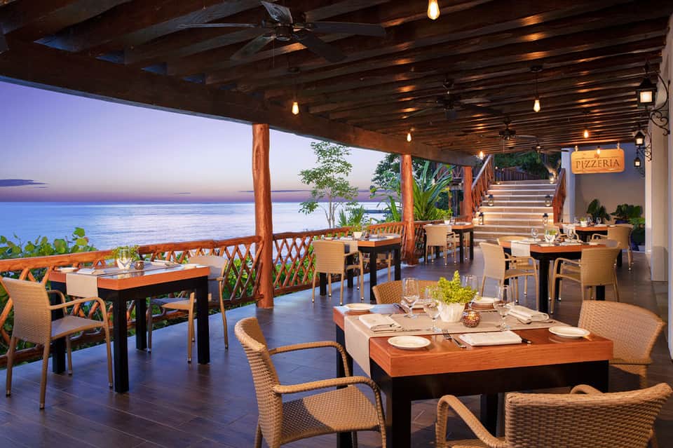 Beachfront pizzeria with wooden beams, ocean sunset view, woven chairs, and rustic dining tables