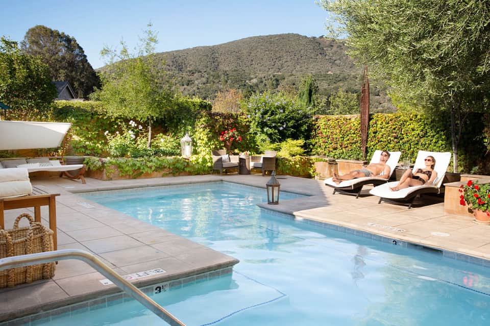 Resort pool area with spa, loungers, green hedges, mountain view, and wooden deck
