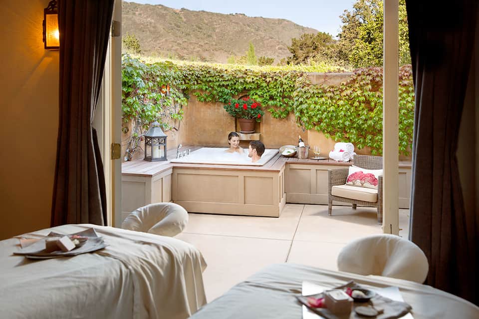 Luxury spa suite with outdoor soaking tub, ivy-covered walls, mountain backdrop, and bedroom lounge