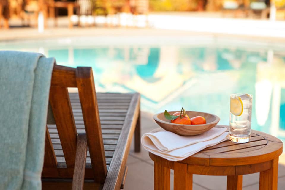 Wooden side table with fruit bowl and water glass beside striped lounger, pool visible in blurred background