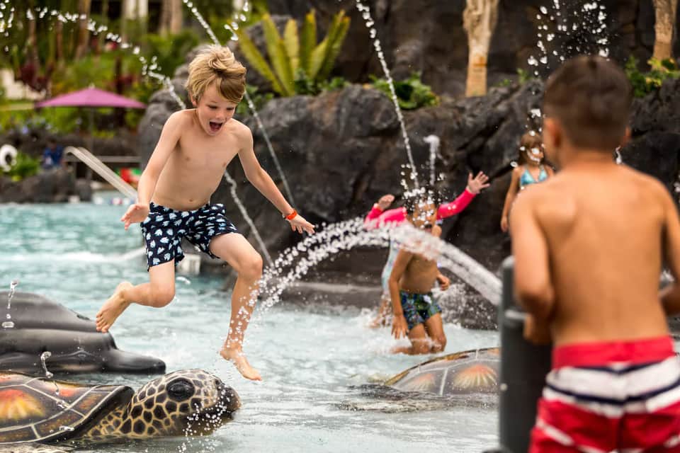 Children playing in resort pool with water fountains and tropical landscaping