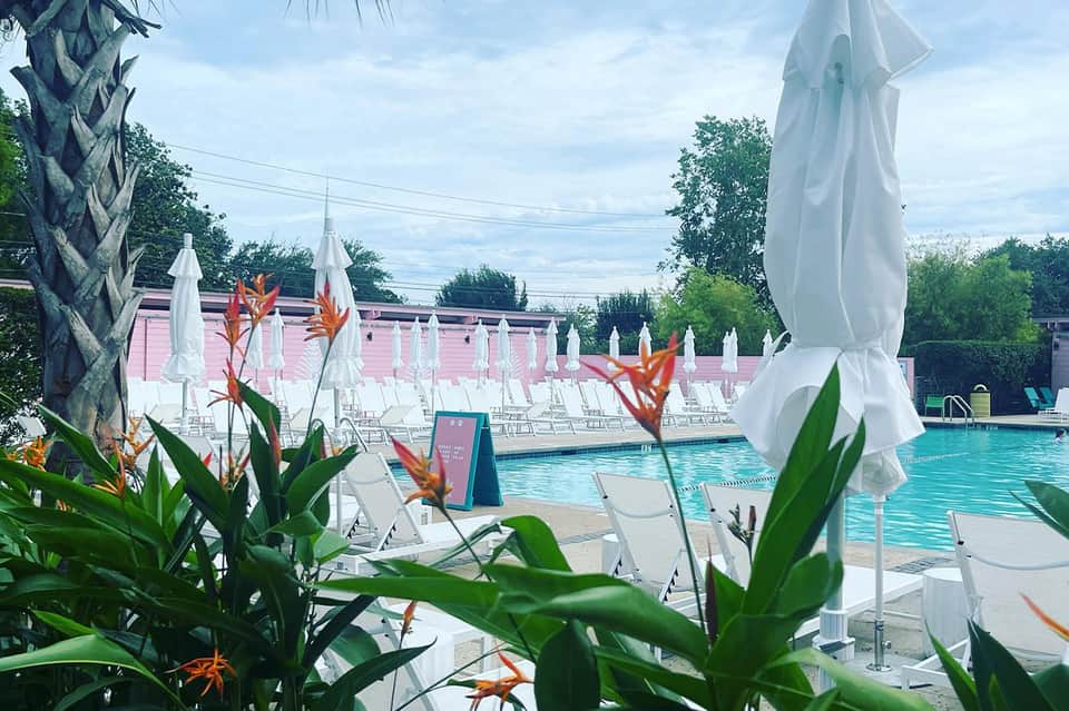 Resort pool with white umbrellas, tropical flowers, pink building, and white lounge chairs in background
