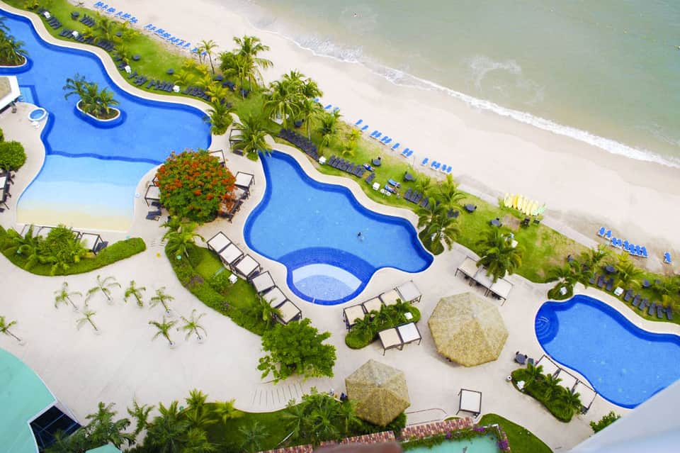 Aerial view of resort pools and beach with palm trees and lounge chairs along sandy shore