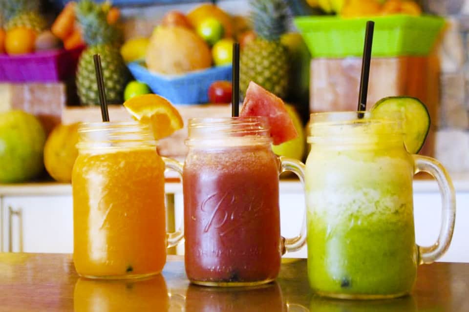 Three colorful tropical fruit drinks in mason jars with straws and fruit garnishes, fresh produce in background