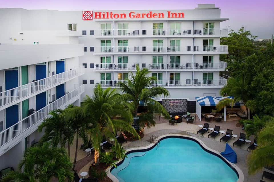 Hilton Garden Inn exterior with white balconies, palm trees, and curved pool at dusk