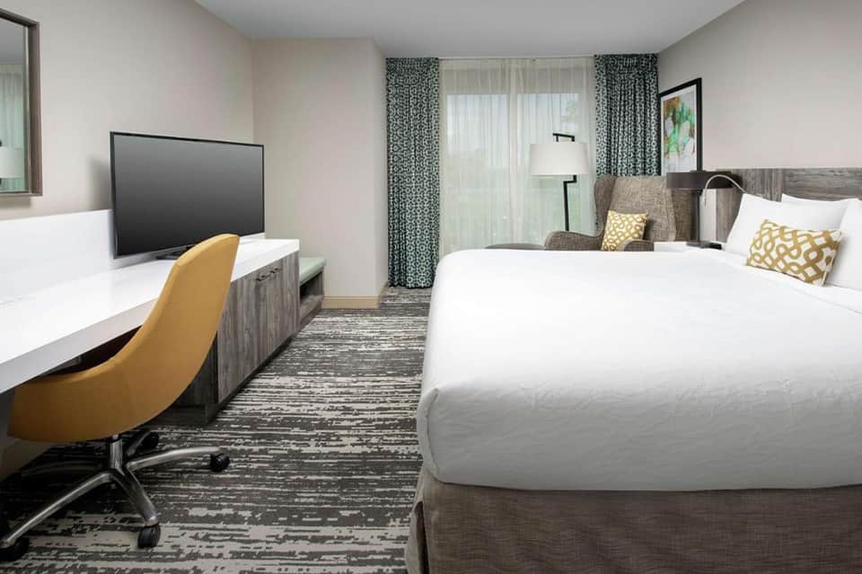 Hotel bedroom with queen bed, work desk with yellow chair, and modern furnishings