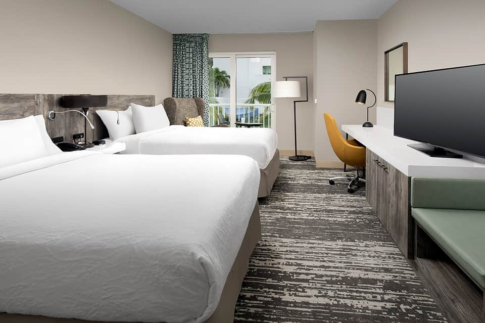Hotel bedroom with two queen beds, ocean-view balcony, modern furnishings, and tropical decor