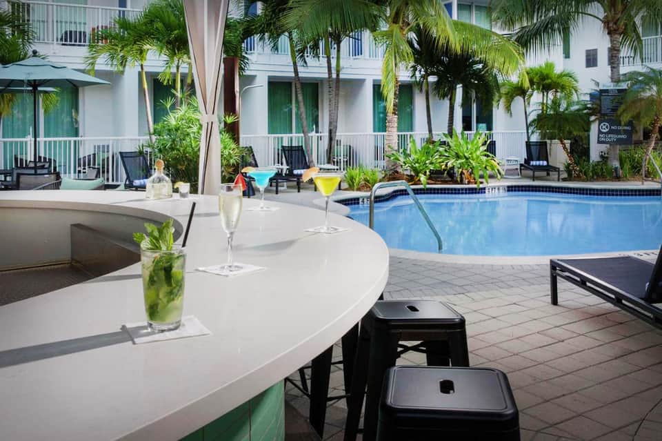 Pool bar area with white tables, cocktails, colorful drinks, and white building with palm trees in background