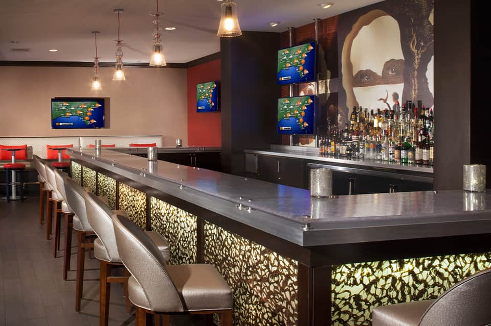 Modern hotel bar with dark wood finishes, illuminated back bar, flat-screen TVs, and geometric patterned panels