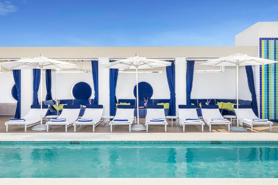 Resort pool deck with white loungers under umbrellas with blue and white cabanas overlooking ocean