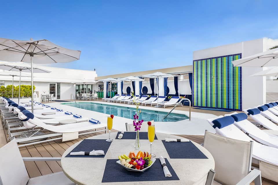 Luxury resort pool with white lounge chairs, blue umbrellas, dining table, and modern architecture