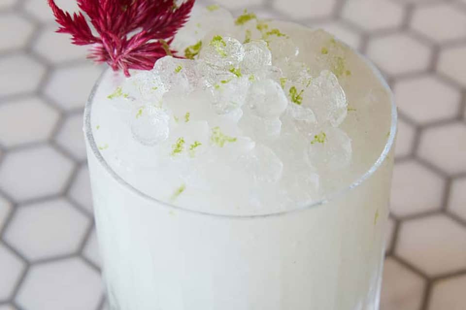 Elegant cocktail with whipped cream, lime zest, and pink flower garnish on hexagonal tile