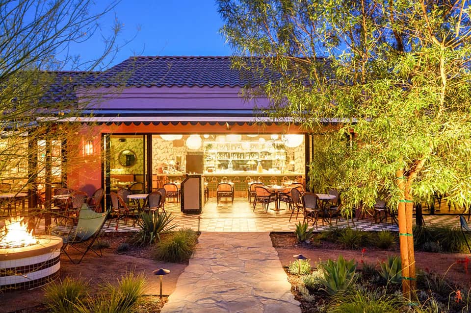Outdoor restaurant pavilion with warm lighting, woven chairs, and garden landscaping at dusk