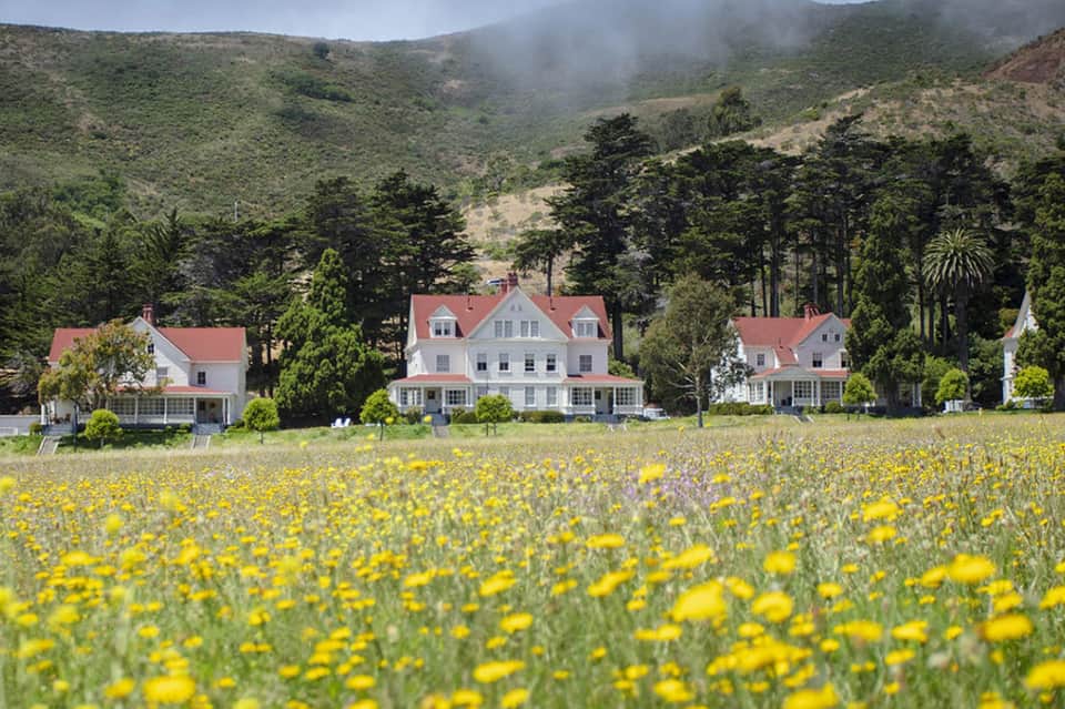 Victorian farmhouse with red roof surrounded by yellow wildflower field and forested mountains