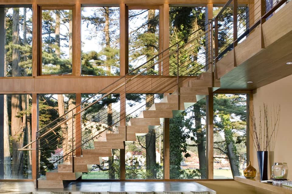 Modern multi-level home interior with floor-to-ceiling windows, wooden stairs, metal railings, and forest views