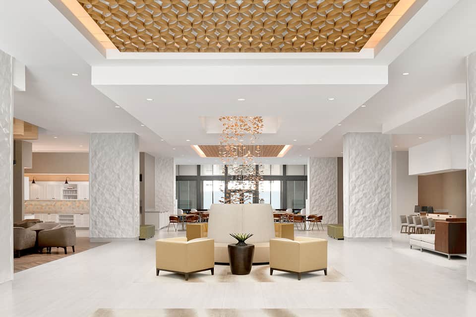 Bright hotel lobby featuring geometric wood ceiling pattern, marble columns, and beige seating areas