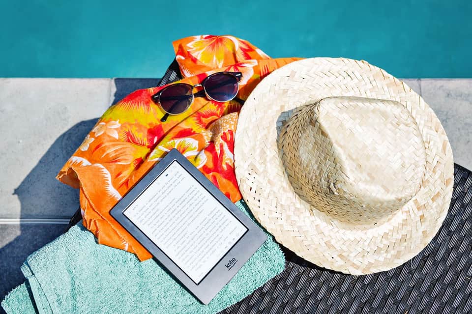 Poolside beach items including colorful sarong, straw hat, sunglasses, towel, and e-reader against turquoise water